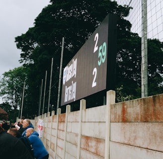 A large digital scoreboard at an outdoor sports venue displays a score of 2-2. Spectators are gathered, observing the event with interest. The scene is enclosed by a tall metal fence and surrounded by lush green trees, indicating a natural setting. Overcast skies add a muted tone to the environment.