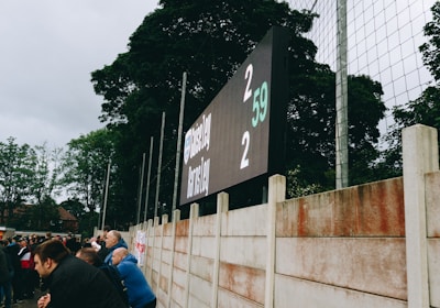 A large digital scoreboard at an outdoor sports venue displays a score of 2-2. Spectators are gathered, observing the event with interest. The scene is enclosed by a tall metal fence and surrounded by lush green trees, indicating a natural setting. Overcast skies add a muted tone to the environment.