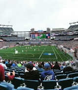 A large sports stadium filled with spectators. The field is green with white markings, and players in red and white uniforms are actively engaged in a game. The stands are packed with people wearing various colored clothing, and some sections of the crowd are more densely populated. Digital screens and advertisements are visible around the stadium, adding to the vibrant atmosphere.