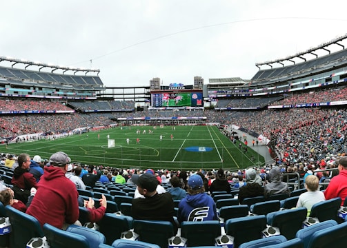 A large sports stadium filled with spectators. The field is green with white markings, and players in red and white uniforms are actively engaged in a game. The stands are packed with people wearing various colored clothing, and some sections of the crowd are more densely populated. Digital screens and advertisements are visible around the stadium, adding to the vibrant atmosphere.
