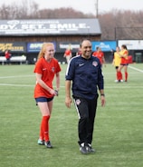 A group of people are on a soccer field, with some wearing sports attire. In the foreground, a person in red sports clothing is walking alongside someone in a dark tracksuit. Other people are visible in the background, some wearing yellow jerseys. A building with advertising banners, including a prominent sign reading 'Ready, Set, Smile,' is in the background.