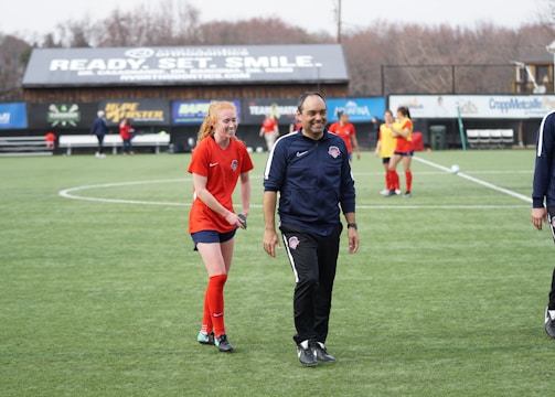 A group of people are on a soccer field, with some wearing sports attire. In the foreground, a person in red sports clothing is walking alongside someone in a dark tracksuit. Other people are visible in the background, some wearing yellow jerseys. A building with advertising banners, including a prominent sign reading 'Ready, Set, Smile,' is in the background.