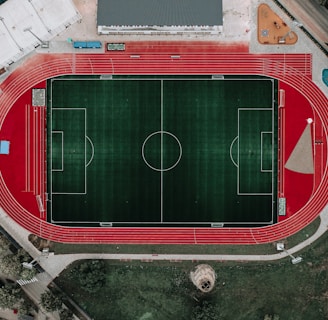 An aerial view of a large sports complex featuring a central green football field surrounded by a red running track. Adjacent to the track, there is a parking area, buildings, and pathways. Nearby, there are additional sports courts in different colors, including a blue swimming pool and orange basketball courts.