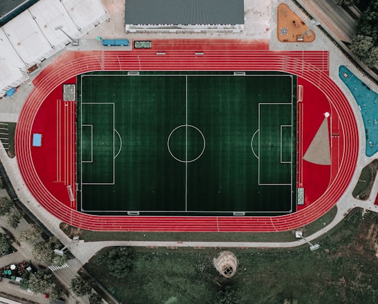 An aerial view of a large sports complex featuring a central green football field surrounded by a red running track. Adjacent to the track, there is a parking area, buildings, and pathways. Nearby, there are additional sports courts in different colors, including a blue swimming pool and orange basketball courts.