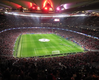 A large, packed stadium with bright lights illuminates a green soccer field. The scene is vibrant with a massive audience surrounding the pitch, visibly excited and engaged. The center of the field displays a prominent circular logo, suggesting a major sporting event. Players and staff can be seen on the field, likely preparing for a match.