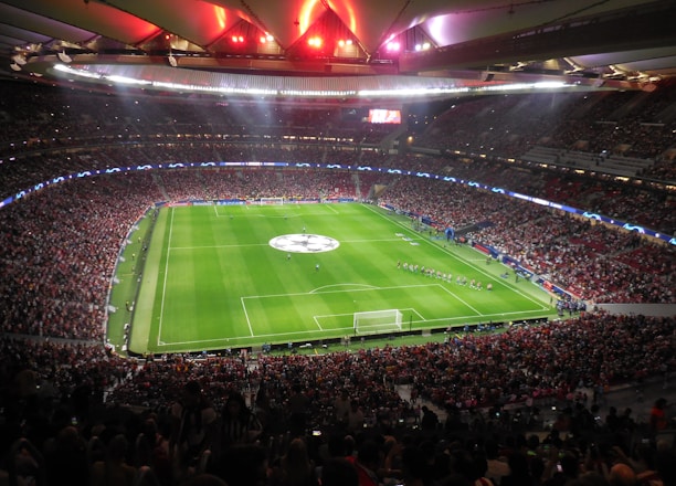 A large, packed stadium with bright lights illuminates a green soccer field. The scene is vibrant with a massive audience surrounding the pitch, visibly excited and engaged. The center of the field displays a prominent circular logo, suggesting a major sporting event. Players and staff can be seen on the field, likely preparing for a match.