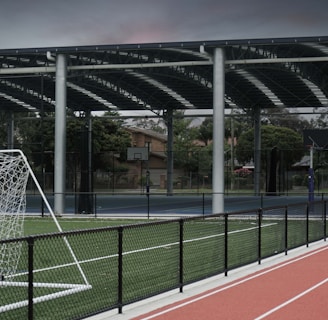 An outdoor sports facility featuring a running track and a soccer goal. The track is red, surrounded by a black metal fence, and the field is covered with artificial green turf. In the background, a basketball court is visible under a covered roof with metal supports. The sky is overcast, adding a moody atmosphere.