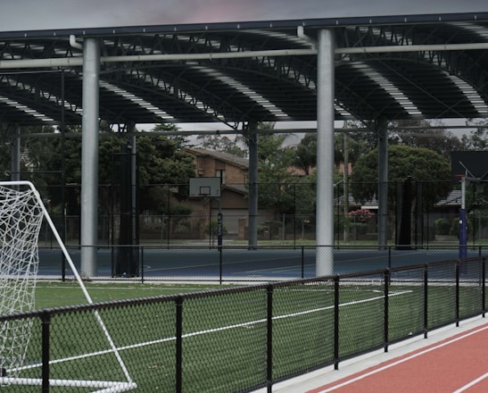 An outdoor sports facility featuring a running track and a soccer goal. The track is red, surrounded by a black metal fence, and the field is covered with artificial green turf. In the background, a basketball court is visible under a covered roof with metal supports. The sky is overcast, adding a moody atmosphere.