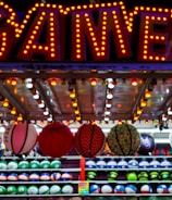 A brightly lit carnival game stall with a large 'GAMES' sign at the top surrounded by red and yellow lights. Various sports balls, including basketballs and soccer balls, are hanging from the ceiling and neatly arranged on shelves below.