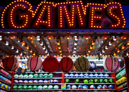A brightly lit carnival game stall with a large 'GAMES' sign at the top surrounded by red and yellow lights. Various sports balls, including basketballs and soccer balls, are hanging from the ceiling and neatly arranged on shelves below.