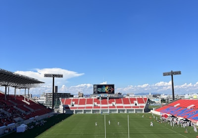 A large sports stadium with red and blue seating overlooks a green field with a goalpost at the center. A few people are scattered across the field, with some tents and a gathering of individuals on the right side. The sky is clear and blue with some clouds visible in the distance. A large screen or scoreboard is situated at the far end of the field.