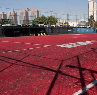 A sports court with a red playing surface and white lines, surrounded by a black mesh fence. In the background, tall buildings are visible, along with some trees and a few yellow chairs positioned near the fence.