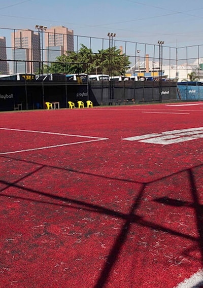 A sports court with a red playing surface and white lines, surrounded by a black mesh fence. In the background, tall buildings are visible, along with some trees and a few yellow chairs positioned near the fence.